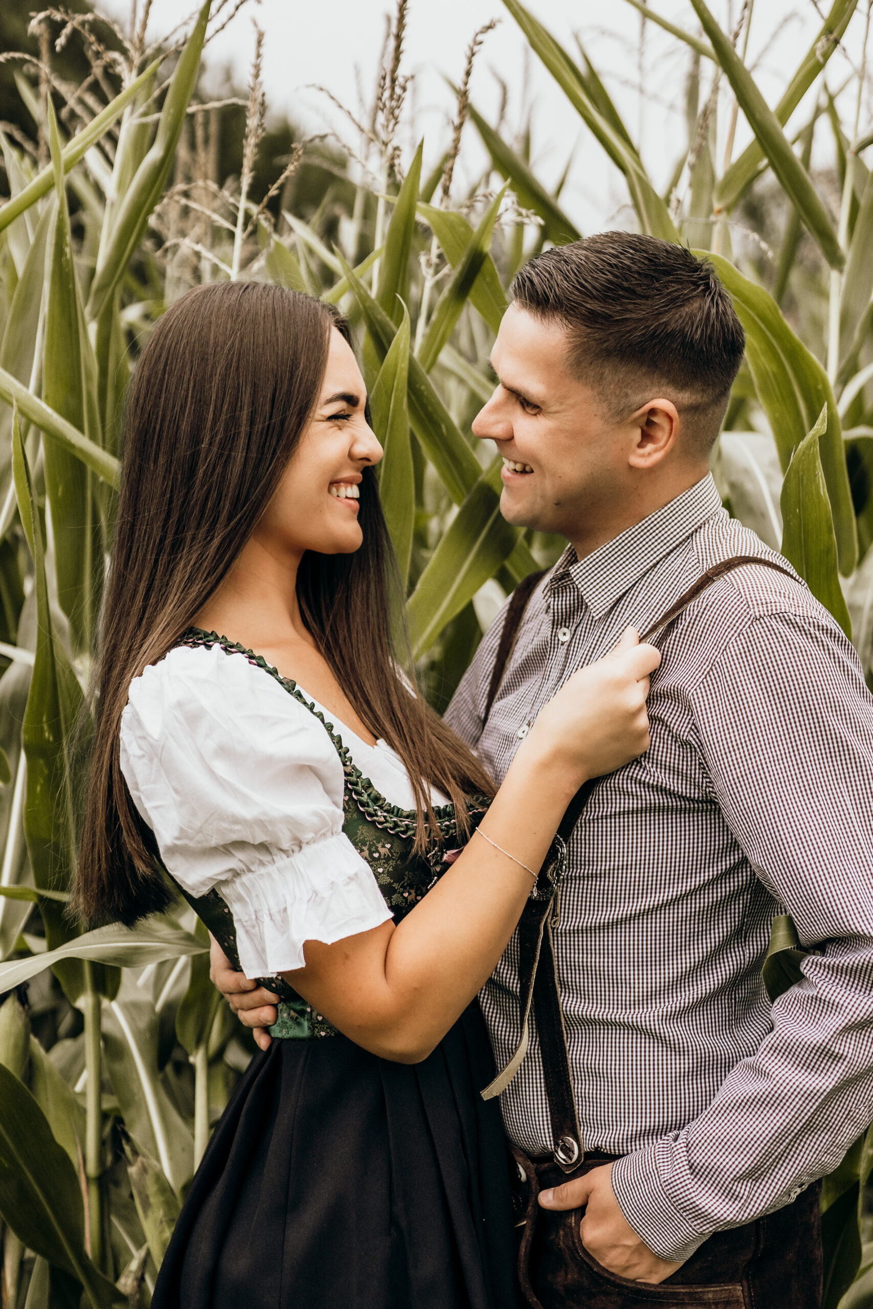 Ein Paar, das einen freudigen Moment während eines Paarshootings in einem Maisfeld teilt, gekleidet in traditionelle Oktoberfest-Kostüme, mit einem Lächeln, das eine tiefe Verbundenheit andeutet.
