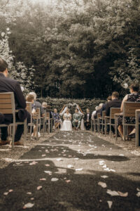 Eine ruhige Hochzeitszeremonie im Freien, bei der die Gäste auf beiden Seiten eines mit Rosenblättern übersäten Ganges sitzen und zu einer Braut und einem Bräutigam im Vordergrund führen, die von üppigem Grün umgeben sind und eine ruhige, intime Atmosphäre teilen